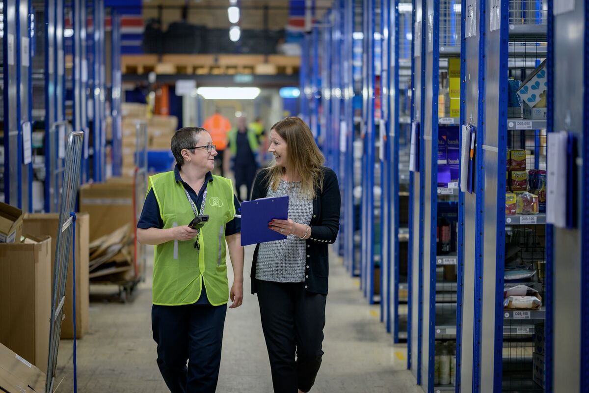 Two women, one with a scanner and the other with a notebook, walking between warehouse racks.