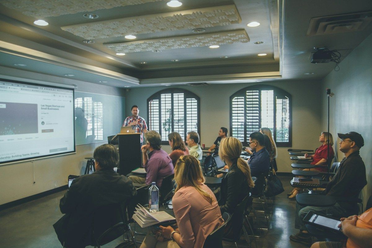 Adult participants listening to an instructor in a training room with a presentation screen.